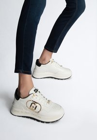 Sneakers in pelle bianca con tallone nero, dettagli dorati e motivi testurizzati laterali; suola in gomma; iscrizione "FATTI PER FAR SENTIRE BENE".