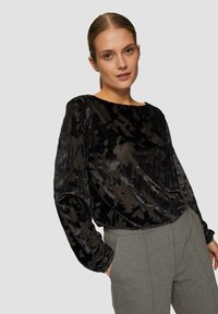 Zwarte fluwelen blouse met een abstract bloemmotief, lange mouwen met geplooide manchetten en een brede ronde hals. Gecombineerd met grijze broeken.