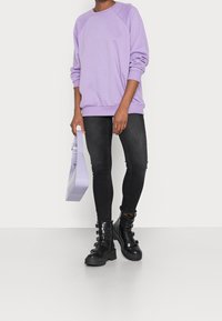 Fialová nadměrná mikina, černé skinny džíny, černé robustní boty se sponou a levandulová texturovaná kabelka. Jednoduchý design a tlumené barvy.