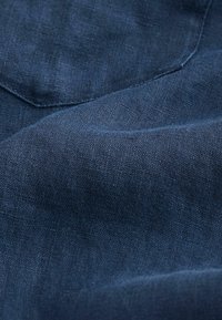 Gros plan d’un tissu texturé bleu foncé avec des coutures visibles et un coin de poche, montrant le tissage et les plis du tissu.
