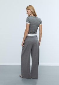 Graues, tailliertes T-Shirt mit weißen Bündchen, kombiniert mit weit geschnittenen grauen Hosen, die eine glatte Textur und Knopfdetails an der Taille aufweisen.