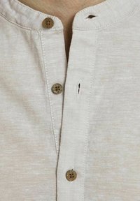 Béžová lenová košile se třemi hnědými knoflíky, z nichž jeden je rozepnutý u krku, což odhaluje část hrudníku nositele.