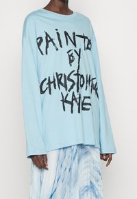 Helles blaues Langarm-T-Shirt mit fettem schwarzen Text, der "PAINTED BY CHRISTOPHER KANE" sagt. Weicher Stoff mit lässiger Passform.