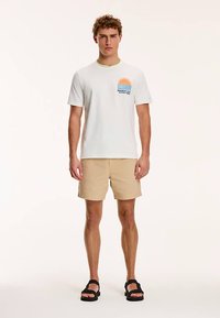 Witte katoenen T-shirt met een kleurrijke zonafbeelding op de borst, gecombineerd met beige shorts en zwarte sandalen. Eenvoudig ontwerp en losse pasvorm.