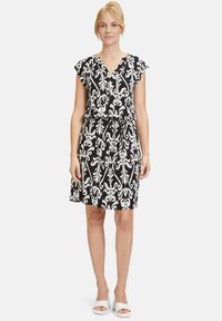 Schwarzes und weißes Kleid mit floralem Muster, kurzen Kappenärmeln, V-Ausschnitt und tailliertem Bund. Weicher Stoff, knielang. Weiße offene Schuhe.