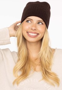 Brown knitted beanie with a ribbed texture, fitted design, and a seamless top. Worn with light-colored, long-sleeve top.