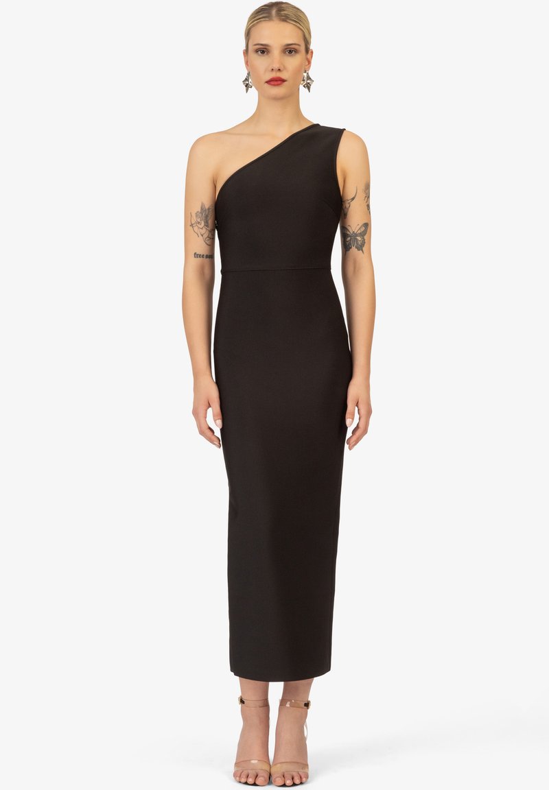 Zwarte midi-jurk met één schouder, een getailleerde silhouette, gladde stof en een rechte zoom. Geaccentueerd met minimalistische lichtbruine hoge hakken.
