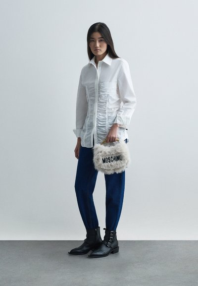 Sieviete, kas ģērbjas baltā ručotā krekla, zila džinsu bikses un melnās potīšu zābaciņos, tur rokās bežīgu, pūkainu Moschino sajūgu ar zelta rokturi.