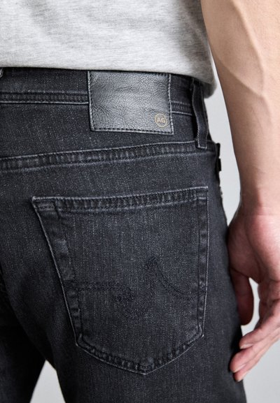 Mörka denimjeans med en slät textur, med en svart läderlapp vid midjan och diskreta sömmar på bakfickorna.