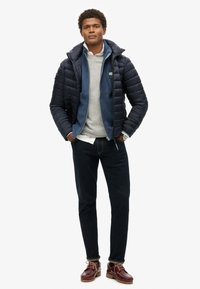 Granatowa kurtka puchowa nałożona na ciemnoniebieski polar, szary sweter, ciemne dżinsy i burgundowe skórzane buty na sznurówki.