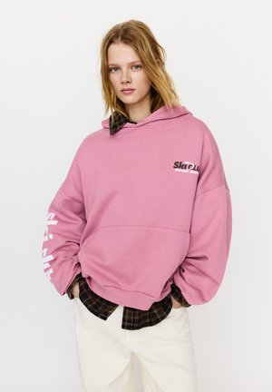 Rosa hoodie med en logotyp på bröstet och text längs ärmarna. Lager-på-lager över en mörk rutig skjorta, matchad med vita byxor.