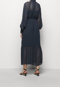 Námorně modré šaty s puntíky, dlouhými balónovými rukávy, vypasovaným pasem a vrstveným lemem. Vyrobené z průsvitné látky; doplněné sandály na řemínky.