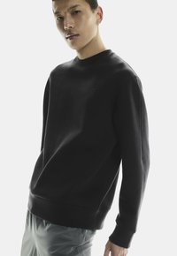 Zwarte crewneck sweatshirt van zacht materiaal; met een subtiele logo aan de voorkant. Gecombineerd met lichtgrijze shorts.