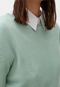Maglione lavorato a maglia verde chiaro con un motivo a diamante testurizzato, scollatura rotonda e una camicia bianca a strati sotto.