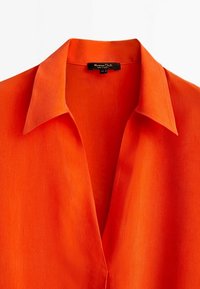 Chemise orange vif à col avec un décolleté en V profond et une petite étiquette noire à l'intérieur du col.