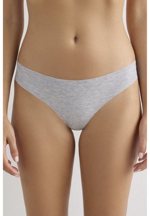 Kobieta w jasno szarym bezszwowym bieliźnie bikini, stojąca z rękami swobodnie opuszczonymi wzdłuż ciała na jednolitym tle.