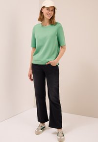Femme portant un t-shirt vert clair à manches courtes, un jean noir à jambes droites, des baskets blanches et vertes, et une casquette blanche, se tenant debout et souriant.