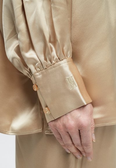 Ruka spočíva pozdĺž tela, oblečená v béžovej saténovej blúzke so zúženým rukávom a manžetou so dvoma gombíkmi a jemným vyšitým logom.