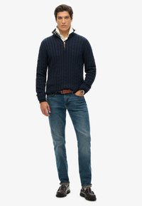 Jonge man die met één hand in zijn broekzak staat, gekleed in een marineblauwe kabelgebreide trui over een crèmekleurig overhemd, blauwe jeans, een bruine riem en donkere instappers.