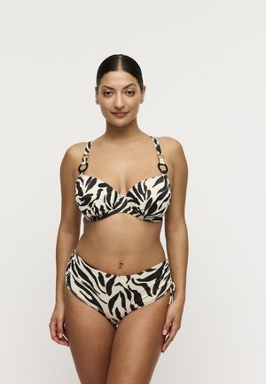 Vrouw staand, naar voren gericht, gekleed in een zwart-witte bikini met zebraprint en verstelbare bandjes, tegen een effen lichte achtergrond.