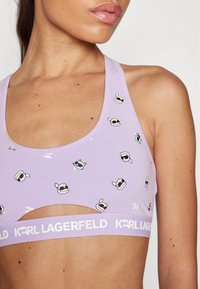 Bra sportivo lilla con design a taglio, realizzato in tessuto elasticizzato. Presenta accenti stampati giocosi e un elastico con logo attorno alla parte inferiore.