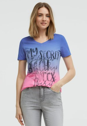 Frau trägt ein blau-rosa verlaufendes T-Shirt mit schwarzem Text und hellgraue Jeans, steht mit einer Hand in der Tasche.