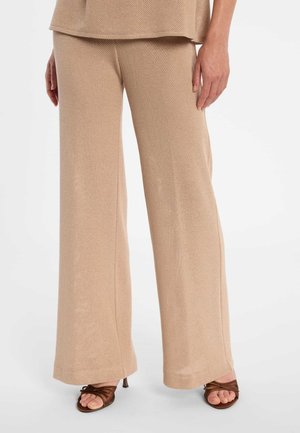 Pantalon large beige texturé porté avec des sandales à talons marron à brides et un haut assorti, présenté de la taille aux pieds sur fond blanc.