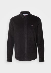 Camisa de pana negra con cuello camisero, mangas largas y cierre frontal de botones. Presenta un pequeño logo en el pecho. Tejido texturizado.