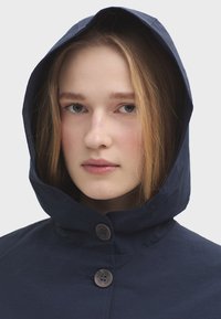 Námornícka mikina s kapucí a vysokým límcem, doplněná o dva hnědé knoflíky a hladkou texturu látky. Jednoduchý design zaměřený na funkčnost.