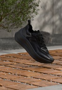 Baskets noires Calvin Klein à semelle épaisse flottant au-dessus d'un sol en carreaux de bois en extérieur, avec un fond de feuillage vert et un mur en béton.