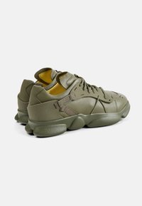 Sneaker verde oliva con tomaia in pelle texturizzata, collare imbottito e suola oversized distintiva. Rivestimento interno giallo e dettaglio della fascia.