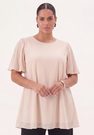 Vrouw met naar achteren gekamd donker haar die een lichtbeige blouse met korte mouwen en zwarte broek draagt, staand met de handen zachtjes in elkaar gelegd.