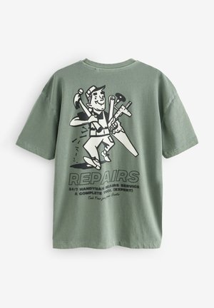 T-shirt in cotone verde oliva con un grafico nero di un tuttofare che tiene degli attrezzi. Il design include la scritta "RIPARAZIONI" e i dettagli del servizio.