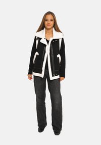 Veste noir et blanc avec un grand col en fourrure, fermeture éclair à l'avant, poches latérales et motifs contrastants ; associée à un jean gris et des chaussures noires.