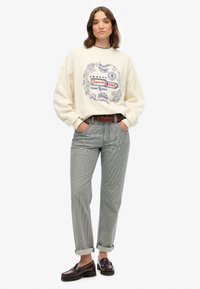 Crème sweatshirt met blauwe graphics en rode accenten, gecombineerd met gestreepte grijze broek en donkerbruine loafers. Ontspannen pasvorm en opgerolde mouwen.