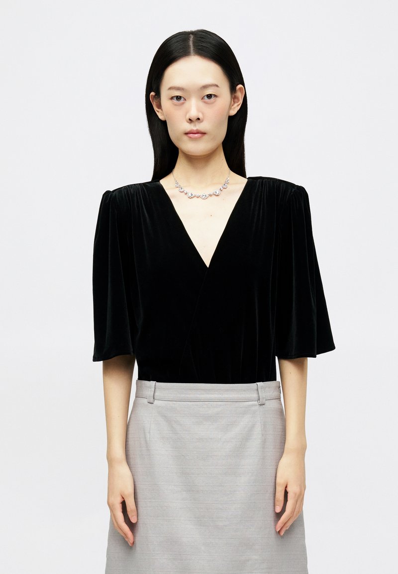 Μαύρη βελουτέ μπλούζα με V-λαιμόκοψη και φουσκωτά μανίκι�α, συνδυασμένη με μια ανοιχτό γκρι φούστα. Απλός ασημένιος κολιέ με λεπτομέρειες. Λεία υφή.