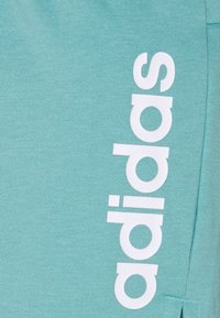Pantaloni in cotone teal presentano un logo "adidas" verticale bianco sul lato sinistro, con una texture liscia e un design a gamba affusolata.