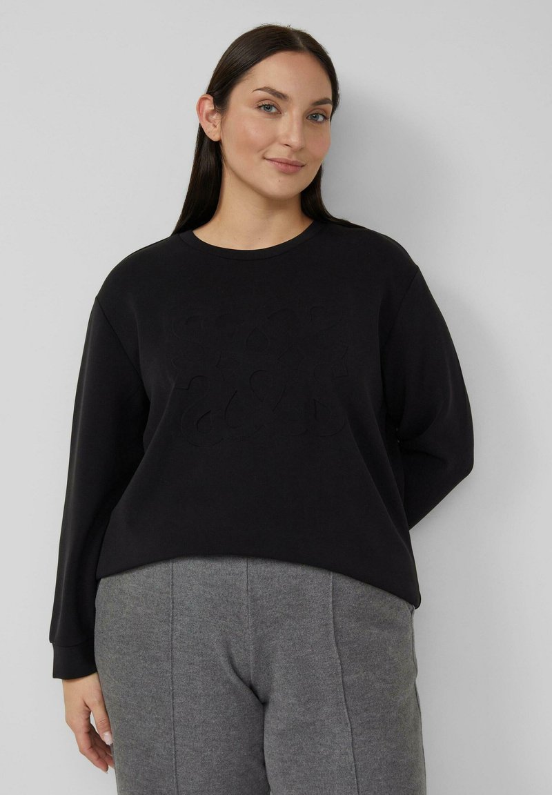 Czarna bluza z miękką teksturą, z wypukłym logo z przodu, okrągłym dekoltem i długimi rękawami. Poniżej widoczne szare spodnie.