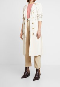 Abrigo crema con botones dorados, cintura ceñida y superpuesto sobre una blusa rosa. Combinado con pantalones beige y botines de tobillo estampados de serpiente marrón.