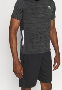 Mustat lyhythihaiset urheilupaidat, joissa on tekstuuripinta, valkoinen Adidas-logo ja kolme valkoista raitaa kummallakin puolella; yhdistettynä mustiin shortseihin.