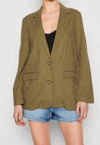 Olivgrön linneblazer med snedskuren krage, två framfickor och två knappar. Matchas med denimbyxor, mjuk textur och avslappnad passform.