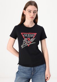 Melns kokvilnas T-krekliņš ar sarkanu trīsstūrainu logo, kurā ir uzraksts "GUESS U.S.A." un kristālu akcenti, kas izsaka "Mīlestība." Ikdienas piegriezums ar īsām piedurknēm.