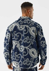 Giubbotto in denim con complessa ricamo paisley bianco, tessuto blu scuro, chiusura con bottoni, due tasche e una vestibilità leggermente sagomata.
