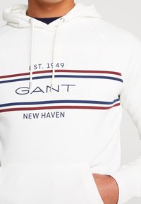Felpa bianca in cotone con tasca frontale. Presenta strisce blu marino e bordeaux, logo "GANT" e testo "NEW HAVEN". Cappuccio con cordino.