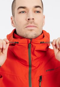 Oranje waterdichte jas met een hoge kraag, verstelbare koorden, voorkant met rits en merkembleem op de borst. Gestructureerde stof.