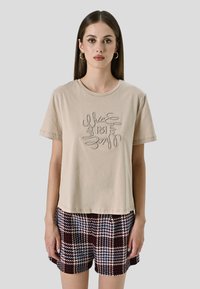 Beige katoenen t-shirt met een ronde hals en korte mouwen, met geborduurde tekst in een circulair ontwerp. Gezamenlijk met geruite shorts.