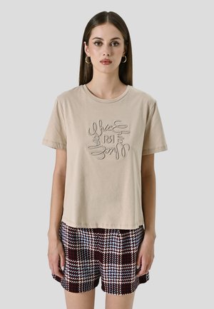 Beige Baumwoll-T-Shirt mit Rundhalsausschnitt und kurzen Ärmeln, das mit gesticktem Text in einem kreisförmigen Design versehen ist. Kombiniert mit karierten Shorts.