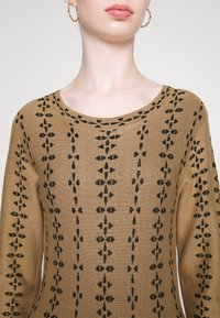 Brązowy sweter dzianinowy z czarnym wzorem kwiatowym, okrągłym dekoltem i długimi rękawami. Tekstura wydaje się gładka z delikatnym połyskiem.