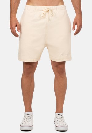 Mężczyzna w beżowych szortach ze ściągaczem i białych trampkach Converse, stojący na jednolitym tle.