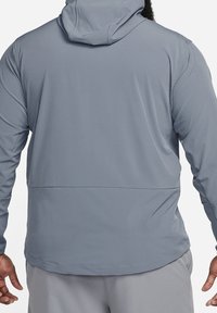 Grijze hooded jack met een gladde textuur, lange mouwen en een met rits afgesloten achterzak. Naaddetails benadrukken het actieve ontwerp.